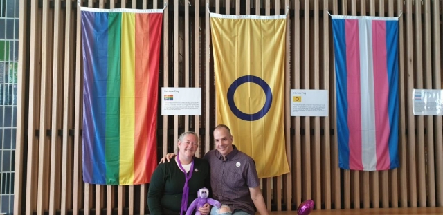 Two people sitting in front of a rainbow flag, intersex flag and transgender flag.