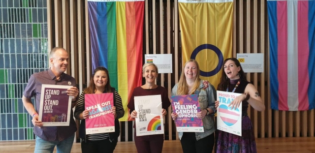 Five people standing in front of rainbow flag, intersex flag and transgender flag holding posters.